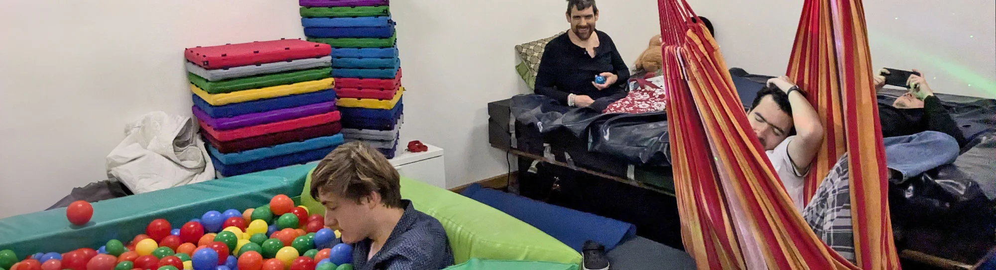 Zwei Bewohner sitzen auf einer Matratze, ein Bewohner sitzt im Bällebad und ein weiterer schaukelt in einer Sitzhängematte
